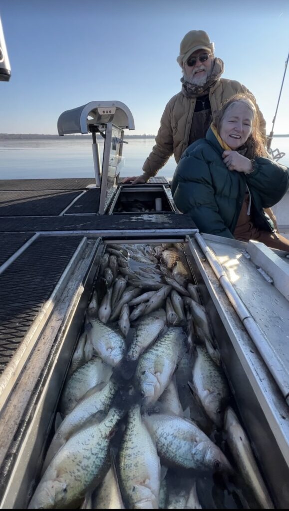 An older man and woman sit on the edge of a boat on a calm body of water. In front of them is a compartment filled with numerous fish. The man wears a beige hat and dark jacket, while the woman wears a green puffy jacket and appears to be smiling.