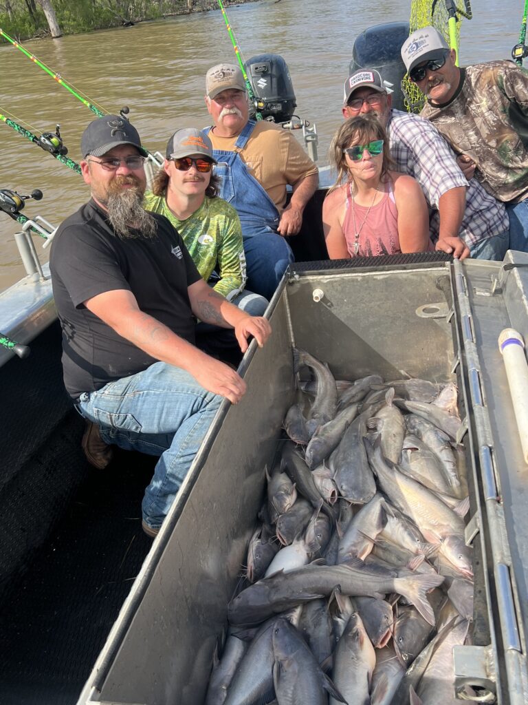 A group of six people poses on a boat holding fishing rods. In the center is a large container filled with numerous fish. The background shows water and some trees. Everyone is smiling and appears to be enjoying a successful fishing trip.