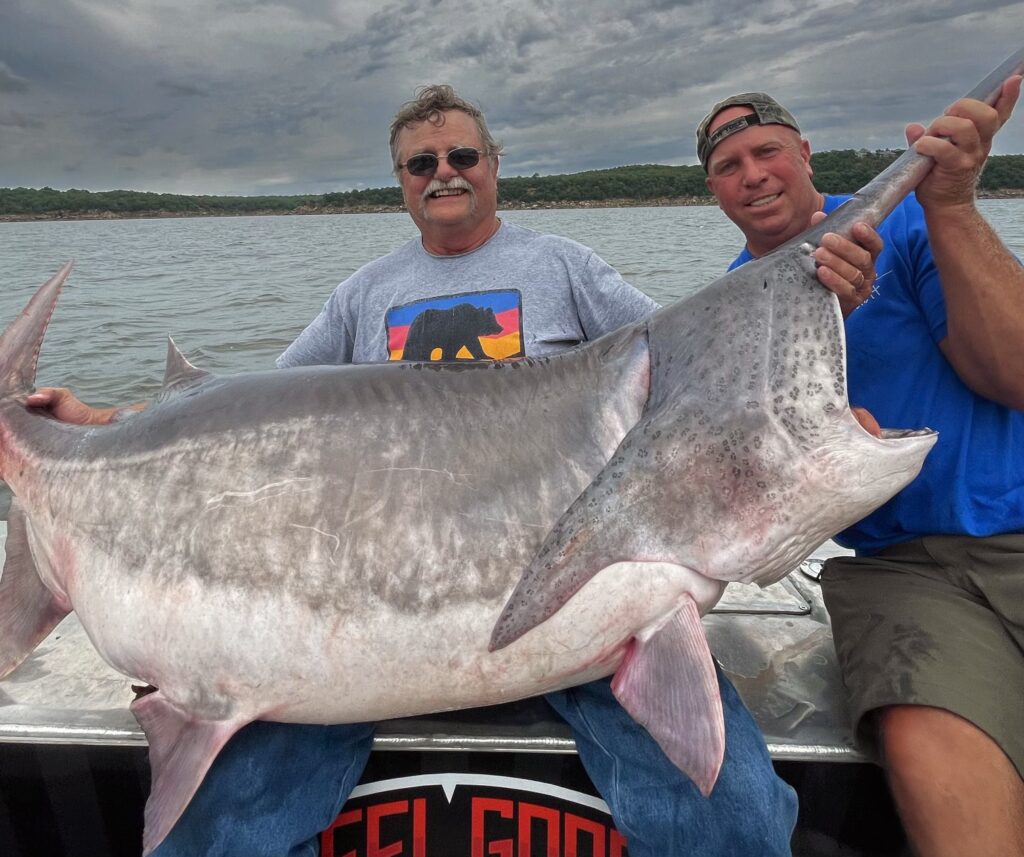 Two men posing proudly on a boat while holding a very large fish. The man on the left is wearing sunglasses and a grey t-shirt with a design on the front, and the man on the right is wearing a blue t-shirt and a cap. Water and cloudy sky are in the background.